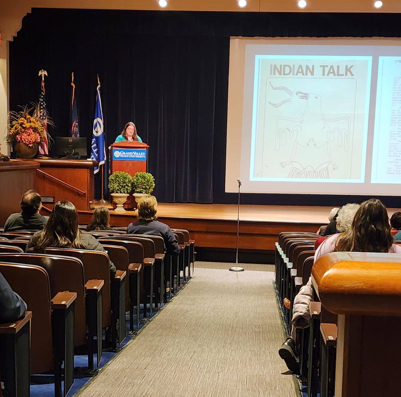 photo of a speaker at a podium on stage in an auditorium with a slide presentation displayed